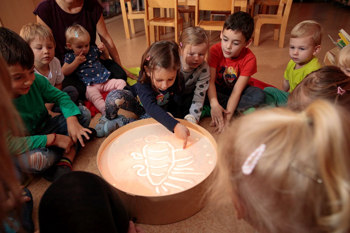 Gruppe von Kindern sitzt im Kreis um einen beleuchteten Sandtisch und schaut einem Mädchen beim Zeichnen zu.