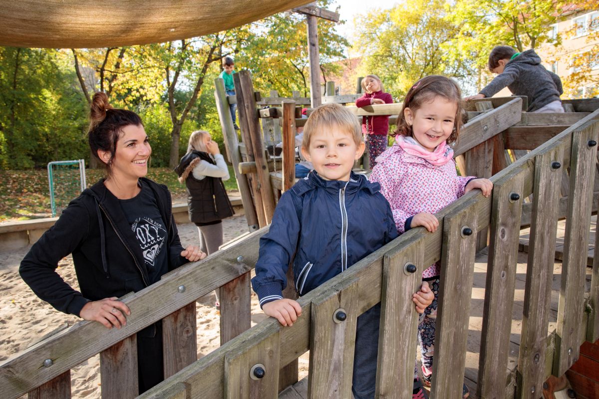 Zwei Kinder stehen mit einer Erzieherin auf einer Holzbrücke im Außengelände und lächeln in die Kamera.