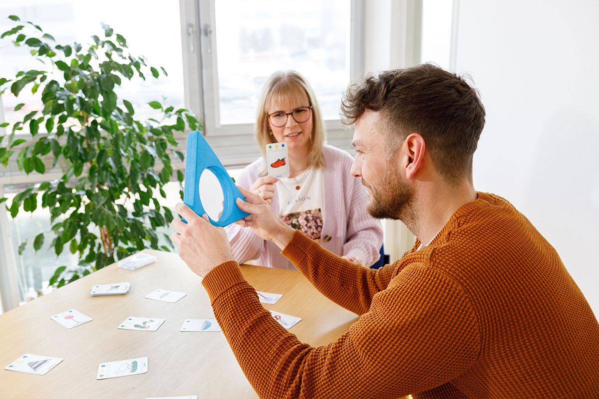 Frau zeigt eine Karte mit einem Bild, Mann betrachtet sie durch eine blaue, dreickige Form; beide sitzen an einem Tisch mit Karten.