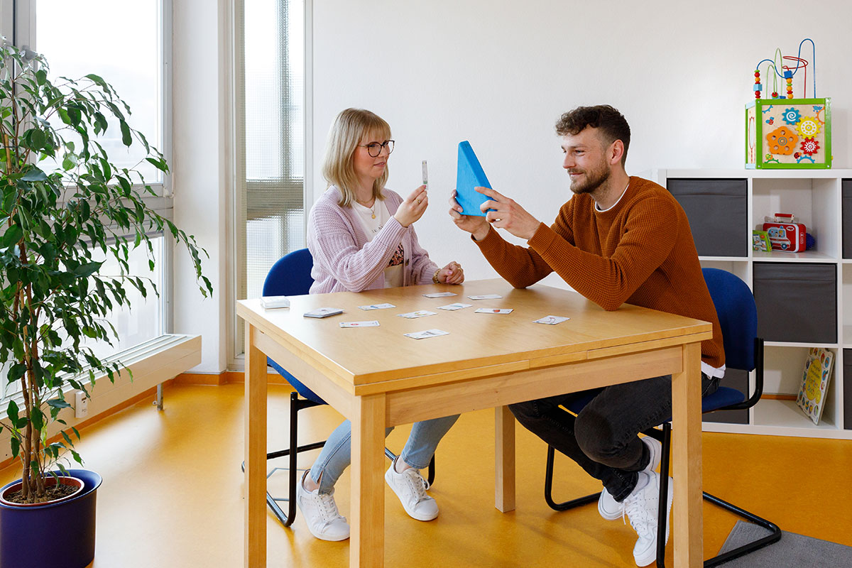 Zwei Erwachsene sitzen an einem Tisch mit Karten und Bildern; sie arbeiten gemeinsam mit einem blauen, dreieckigem Gegenstand im Therapieraum