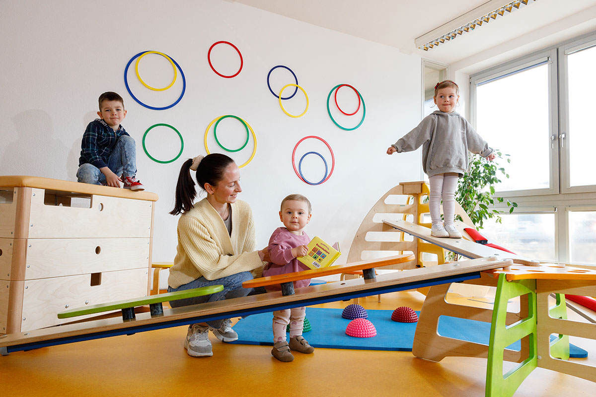 Drei Kinder spielen und balancieren in einem Bewegungsraum, eine Erwachsene fördert dabei spielerisch ein Kleinkind.