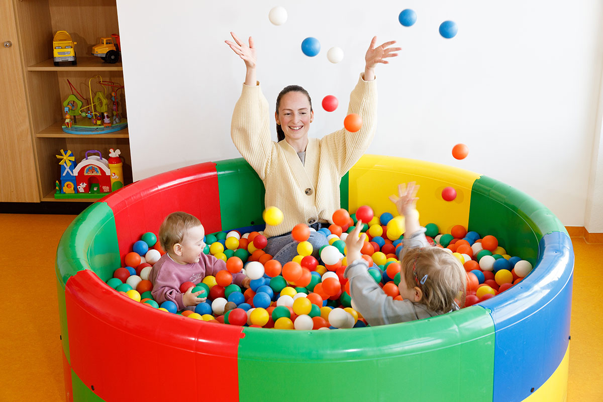 Erwachsene sitzt mit zwei kleinen Kindern in einem bunten Bällebad und wirft fröhlich bunte Bälle in die Luft.